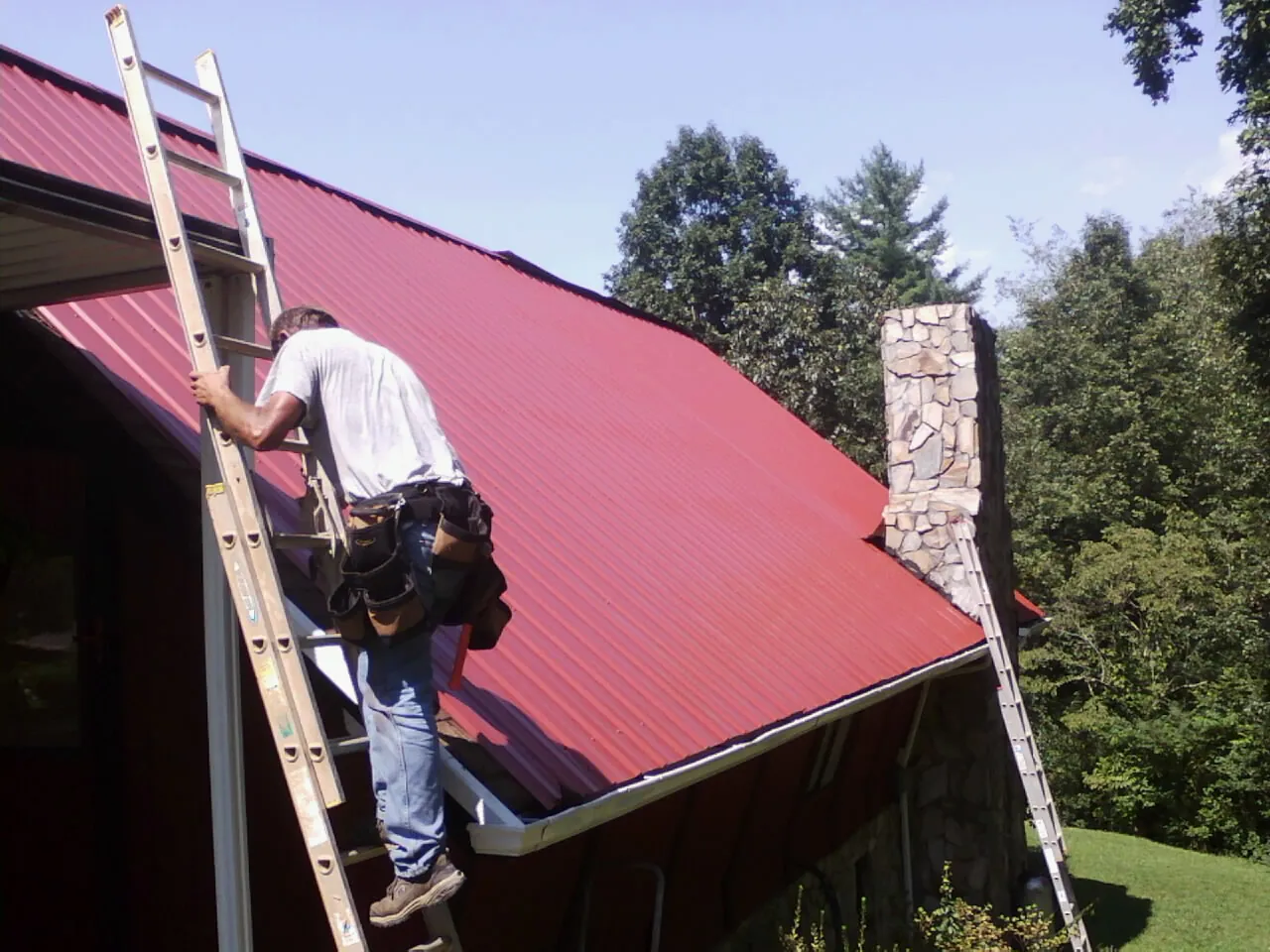 Roofer on a ladder beside a red metal roof and stone chimney — Soffit & Fascia Repair by Roofing Brothers in South Wellfleet